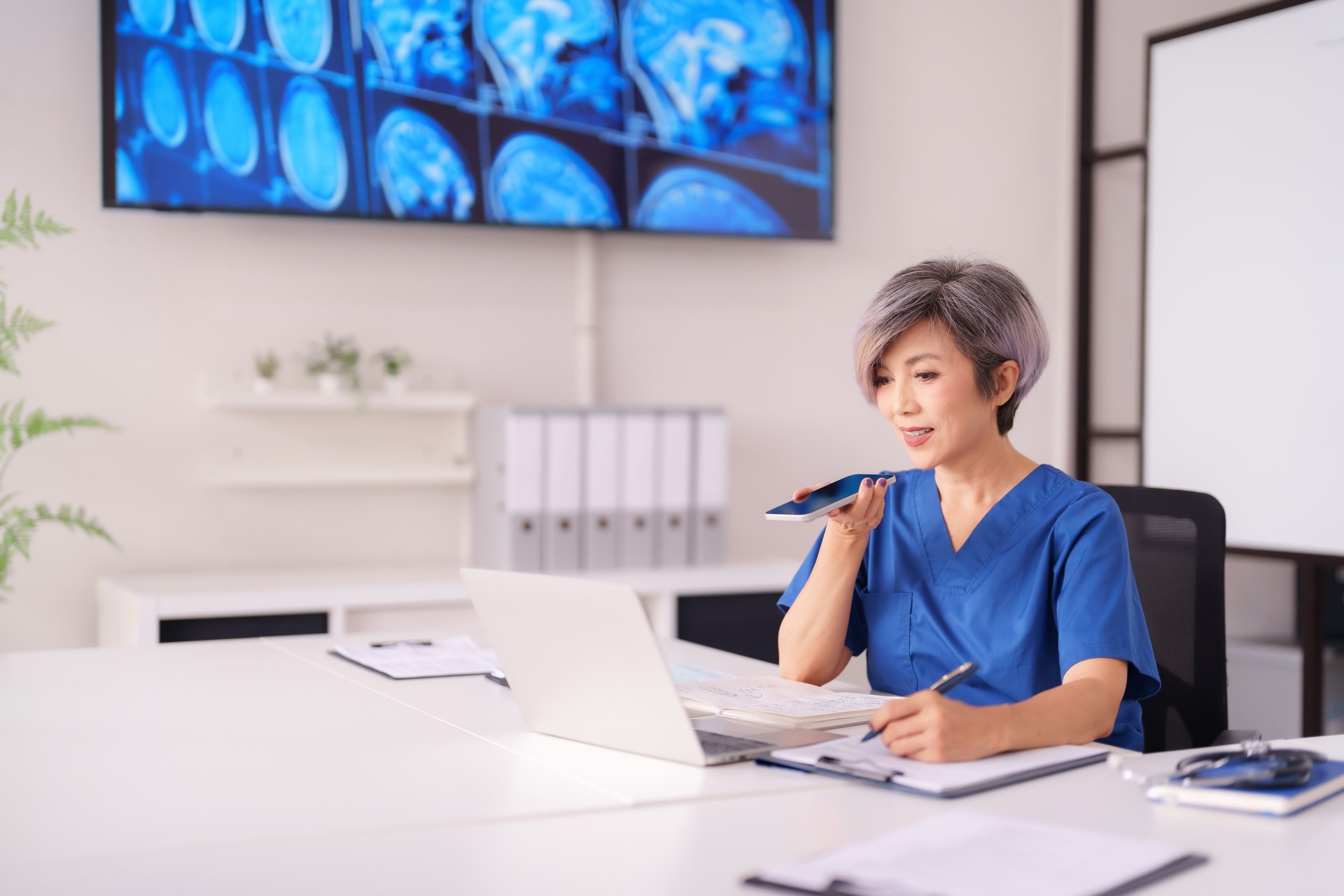 Asian Adult Female Doctor Healthcare Professional Utilizes Smartphone Technology and Laptop for Patient Data Management in Modern Clinic Office