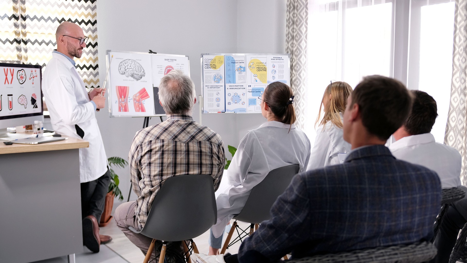 A group of people are sitting on chairs in a room and listening to a man in a white lab coat speak. Doctors attend lectures on medicine and science